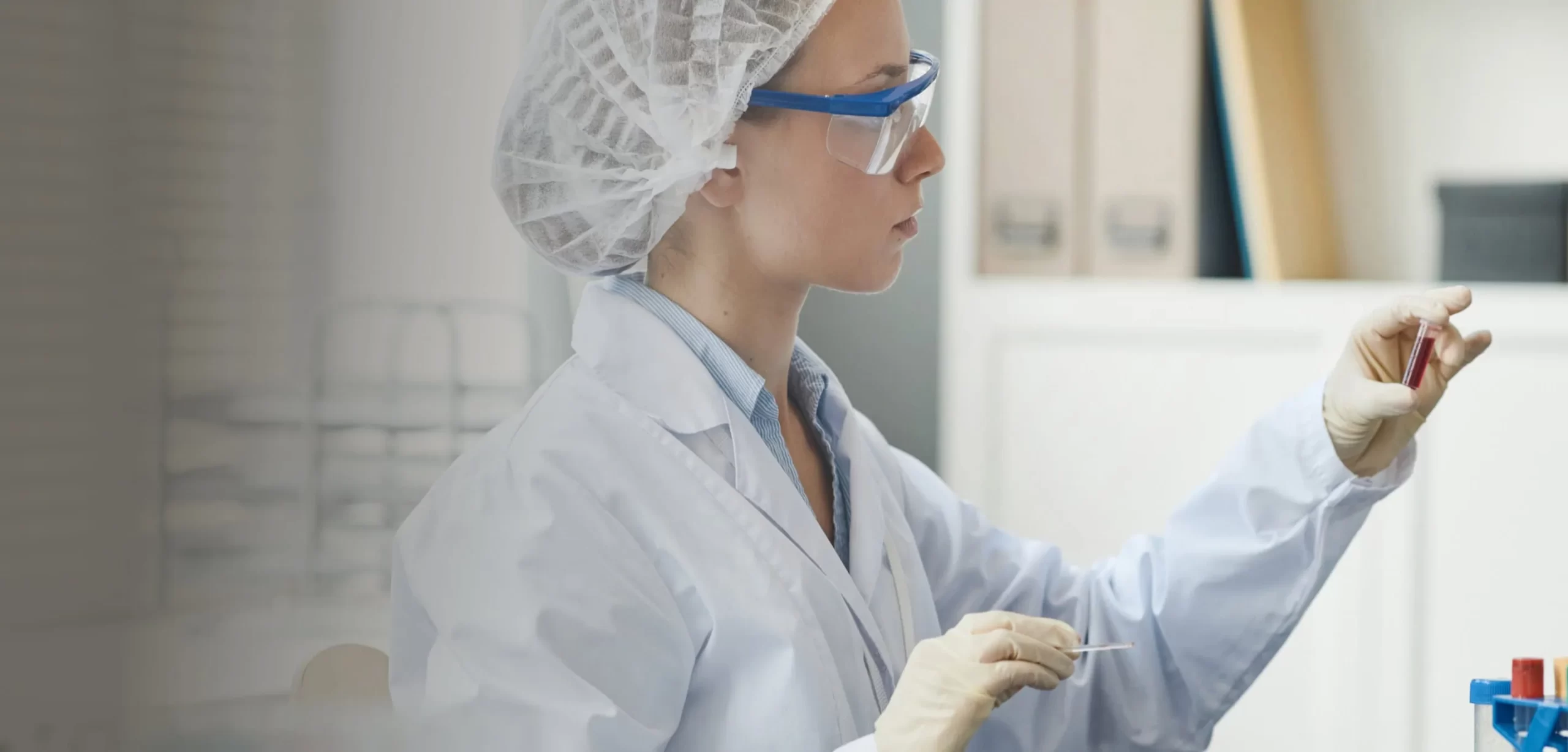 Woman in lab coat and safety goggles examining vial