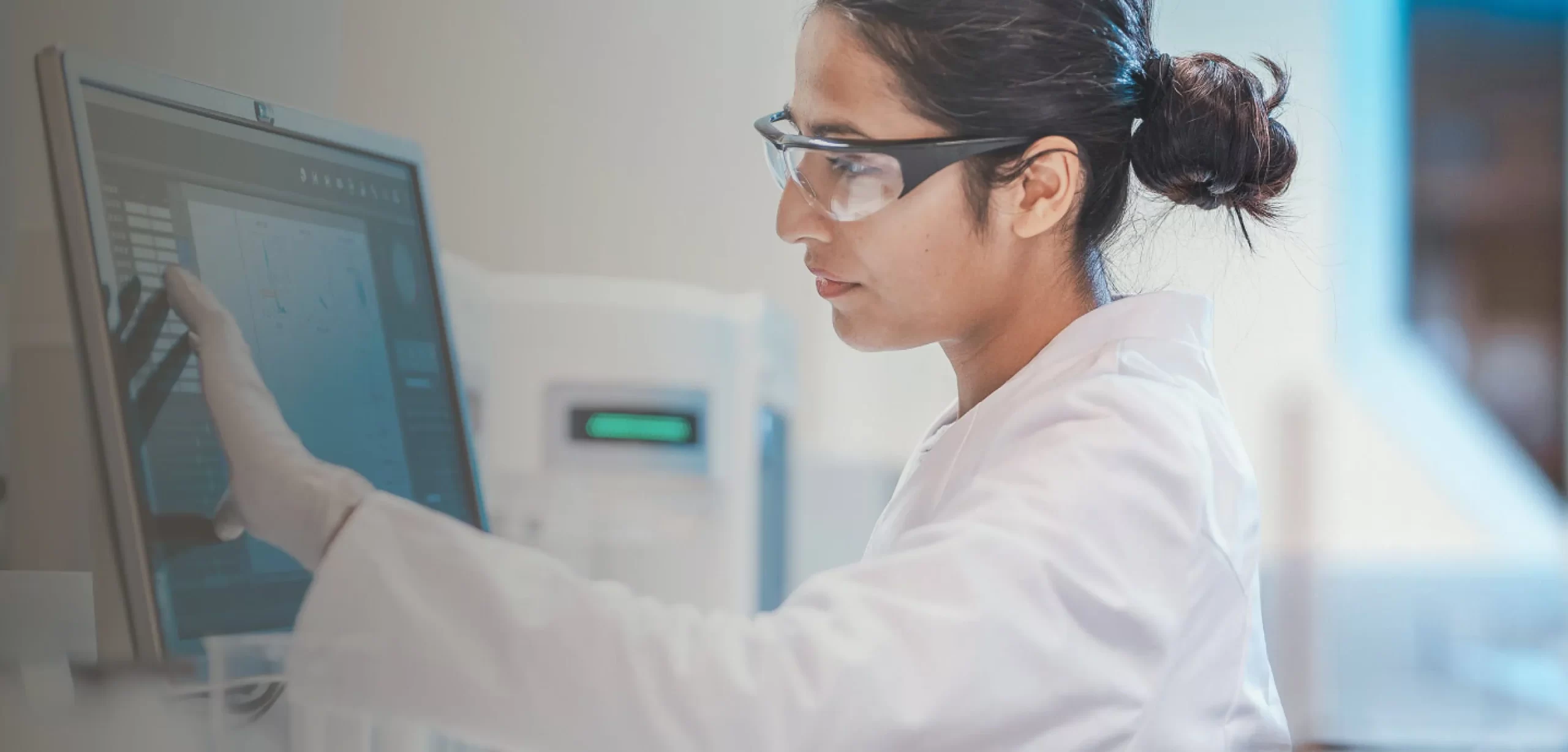 Woman in lab coat and safety googles reviewing touchscreen display